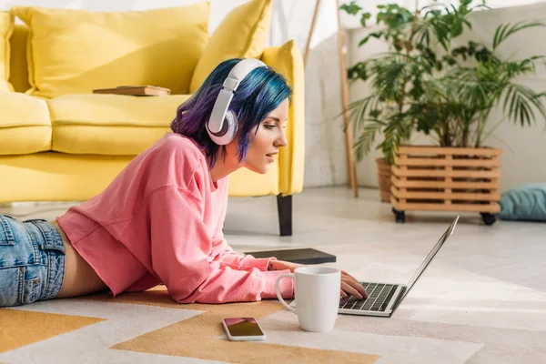 Freelance avec cheveux colorés et écouteurs travaillant avec ordinateur portable près de tasse de thé et smartphone sur le sol dans le salon — Photo de stock