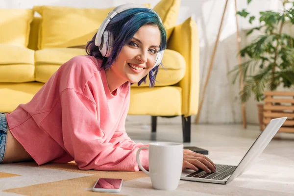 Freelance avec des cheveux colorés dans les écouteurs travaillant avec ordinateur portable près de tasse de thé et smartphone, souriant et regardant la caméra sur le sol — Photo de stock