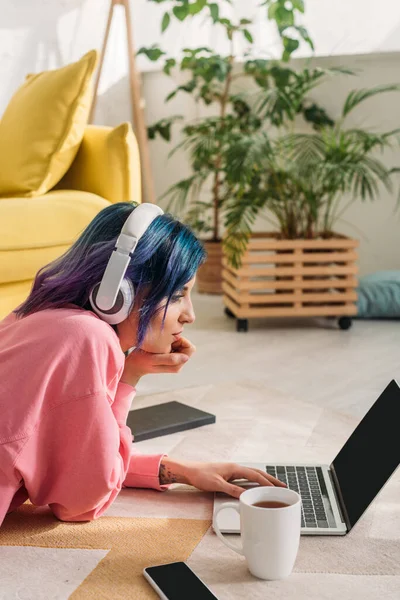 Freelancer com cabelos coloridos e fones de ouvido trabalhando com laptop perto de xícara de chá, notebook e smartphone no chão — Fotografia de Stock
