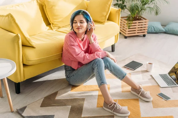 High angle view of freelancer with colorful hair, closed eyes and headphones smiling near sofa on floor — Stock Photo