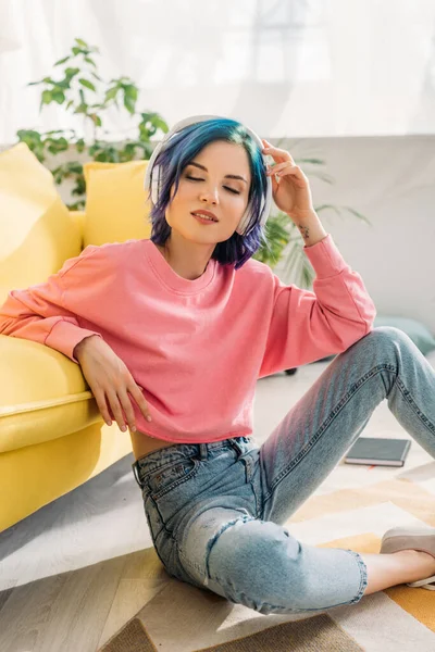 Mujer con el pelo colorido, ojos cerrados y auriculares escuchando música cerca del sofá en el suelo - foto de stock