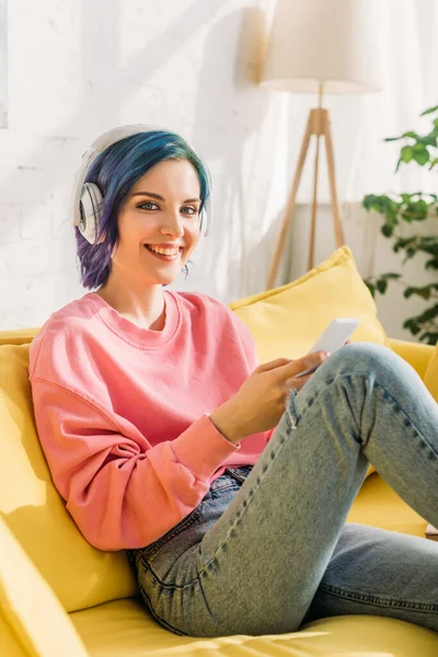 Mujer con el pelo colorido y auriculares sosteniendo teléfono inteligente, sonriendo y mirando a la cámara en el sofá - foto de stock