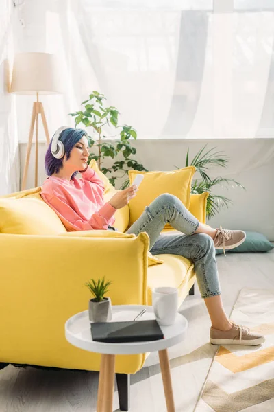 Жінка з барвистим волоссям і навушниками тримає смартфон на дивані біля журнального столика у вітальні — стокове фото