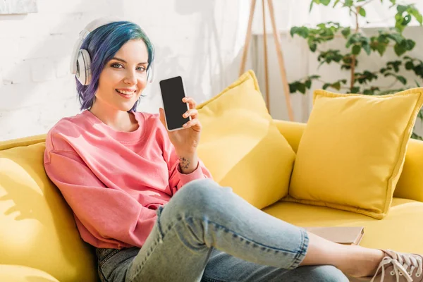 Femme aux cheveux colorés et aux écouteurs souriants, regardant la caméra et montrant un smartphone sur le canapé dans le salon — Photo de stock