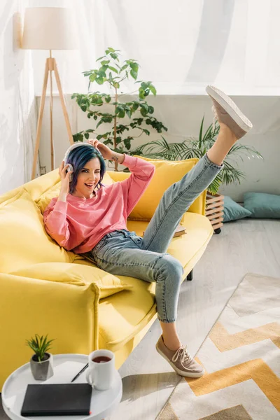 Mulher com cabelo colorido se divertindo com fones de ouvido e perna levantada no sofá na sala de estar — Fotografia de Stock