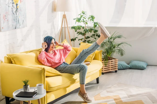 Mujer con el pelo colorido, auriculares y pierna levantada sonriendo en el sofá en la sala de estar - foto de stock