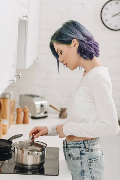 Chica con el pelo colorido preparando comida y tocando la tapa de la sartén cerca de cocina estufa - foto de stock
