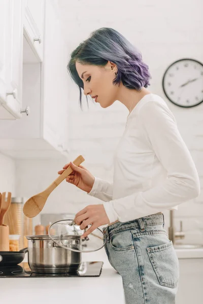 Chica con pelo colorido sosteniendo tapa de la cacerola y espátula cerca de cocina estufa - foto de stock