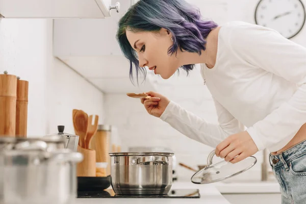 Enfoque selectivo de chica con el pelo colorido preparando comida y sosteniendo la tapa de la sartén con espátula cerca de la cocina estufa - foto de stock