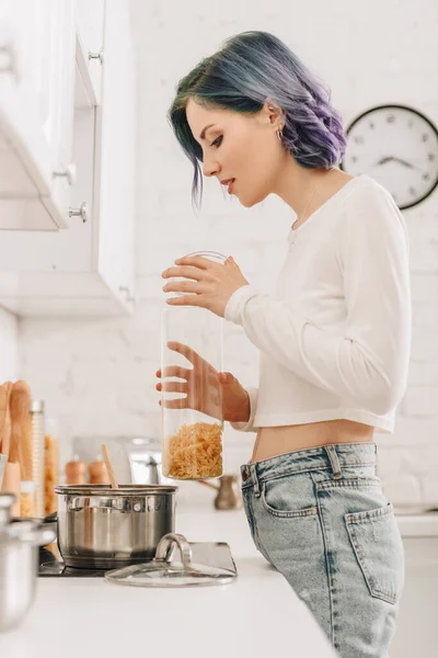 Concentration sélective de la fille avec récipient de tenue de cheveux colorés avec des pâtes près du poêle de cuisine — Photo de stock
