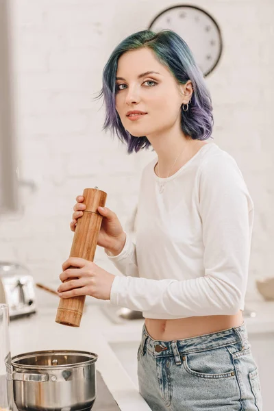 Selective focus of girl with colorful hair smiling and looking at camera with pepper mill above pan in kitchen — Stock Photo