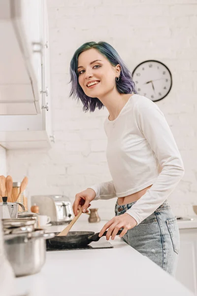 Visão de baixo ângulo da menina com cabelo colorido preparando alimentos com espátula na frigideira, sorrindo e olhando para a câmera na cozinha — Fotografia de Stock