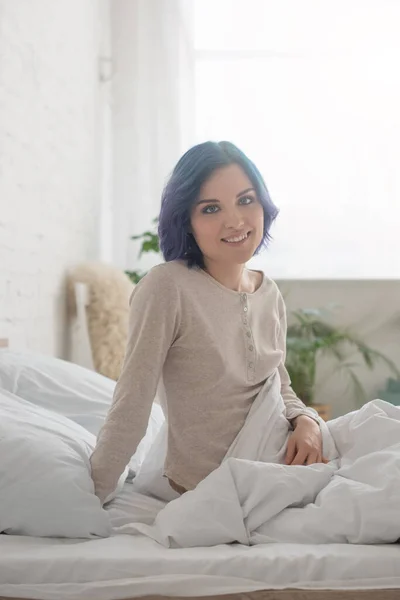 Donna con i capelli colorati sorridente e guardando la fotocamera sul letto in camera da letto — Foto stock