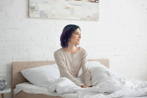 Attractive woman with colorful hair looking away and smiling on bed in bedroom — Stock Photo