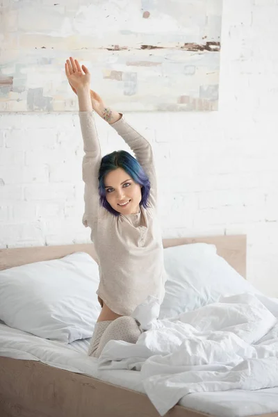 Mulher com cabelos coloridos esticando-se, olhando para a câmera e sorrindo na cama no quarto — Fotografia de Stock