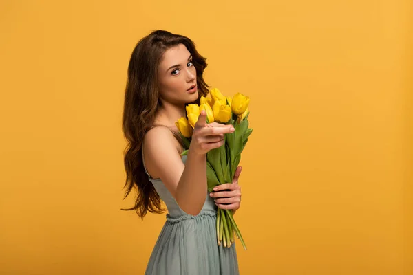 Menina bonita em vestido de primavera apontando e segurando buquê de flores isoladas no amarelo — Fotografia de Stock