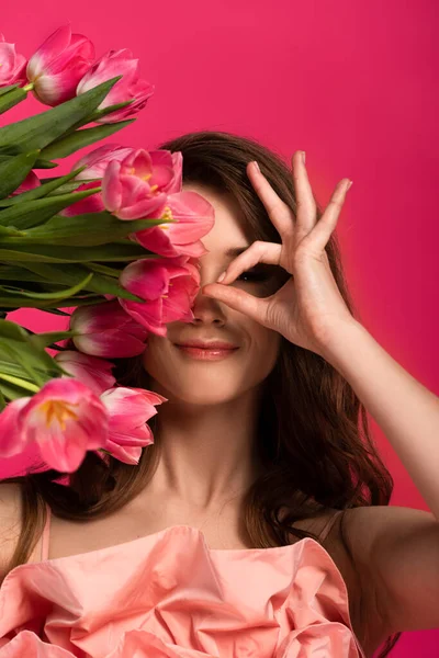 Hermosa chica sonriente con tulipanes de primavera mostrando signo ocular aislado en rosa - foto de stock