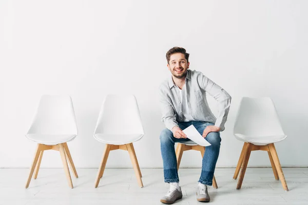 Lächelnder Mann mit Lebenslauf blickt in die Kamera, während er im Büro auf einem Stuhl sitzt — Stockfoto