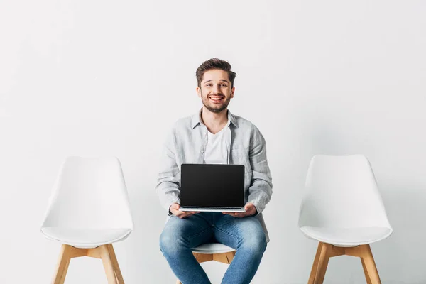 Lächelnder Mitarbeiter blickt in die Kamera, während er Laptop mit leerem Bildschirm auf Stuhl hält — Stockfoto
