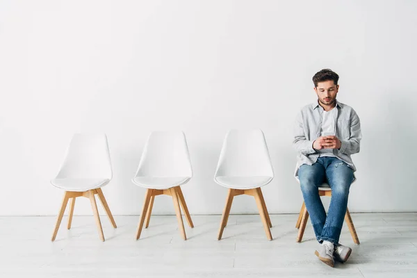 Empleado guapo usando teléfono inteligente mientras está sentado en la silla en la oficina - foto de stock