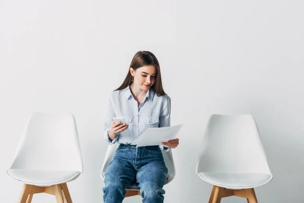 Belle femme utilisant un smartphone et tenant le CV sur la chaise dans le bureau — Photo de stock