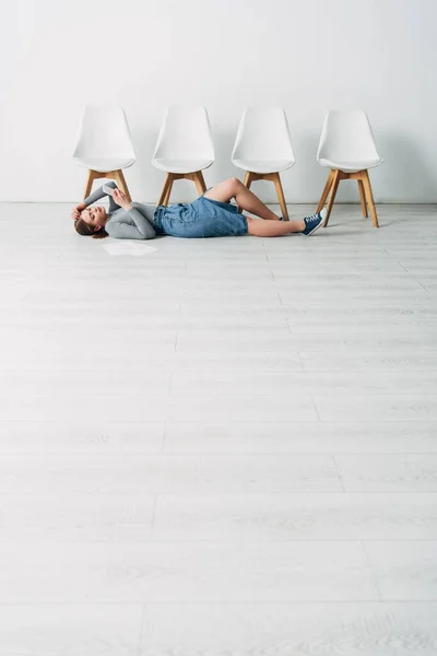 Niedrigwinkel-Ansicht attraktiver Mitarbeiter mit Smartphone, während sie im Büro in der Nähe des Lebenslaufs auf dem Boden liegen — Stockfoto