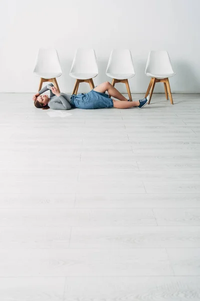 Low angle view of attractive employee using smartphone while lying near resume on floor in office — Stock Photo