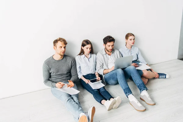 Mujer joven con curriculum vitae mirando a la cámara cerca de los empleados con aparatos en el piso en la oficina - foto de stock