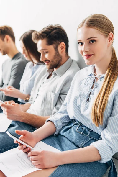 Selective focus of smiling girl with smartphone and resume looking at camera near candidates in office — Stock Photo