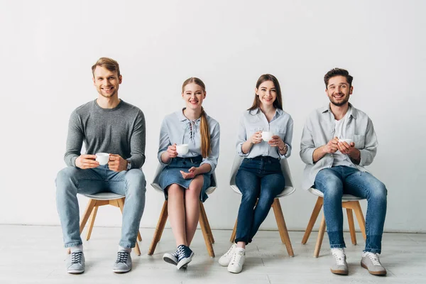 Smiling coworkers with coffee cups smiling at camera on chairs in office — Stock Photo