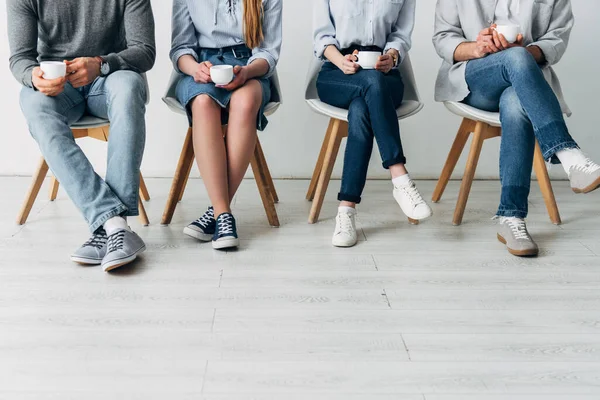 Cropped view of colleagues holding coffee cups on chairs in office — Stock Photo