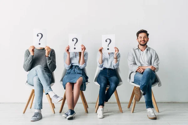 Hombre guapo sonriendo a la cámara cerca de empleados sosteniendo tarjetas con signos de interrogación mientras esperan entrevista de trabajo - foto de stock