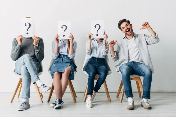 Cheerful man showing thumbs up near candidates with question marks on cards in office — Stock Photo