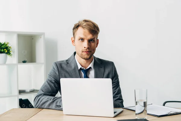 Schöner Personalvermittler schaut in die Kamera neben Laptop und Klemmbrett auf dem Tisch — Stockfoto