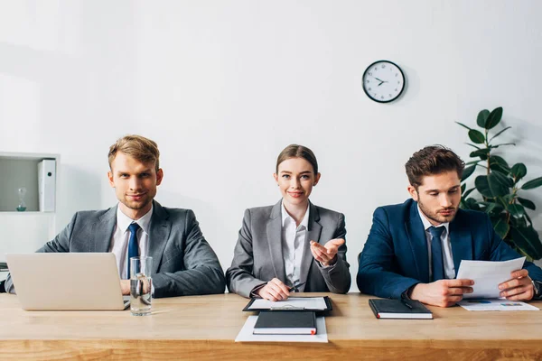 Lächelnde Recruiter mit Papieren und Laptop blicken in die Kamera am Tisch — Stockfoto