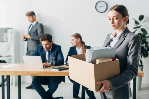 Mujer despedida sosteniendo caja de cartón cerca de colegas que trabajan en la oficina - foto de stock