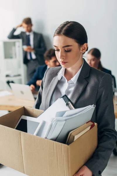 Selective focus of fired woman holding box with documents and digital tablet while colleagues working in office — Stock Photo