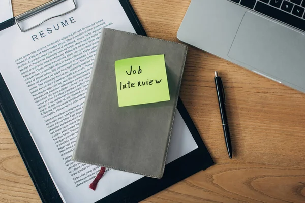 Vista superior del cuaderno con letras de entrevista de trabajo en la nota adhesiva en el curriculum vitae cerca de la computadora portátil en la mesa — Stock Photo