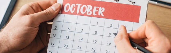 Vista ritagliata dell'uomo che tiene il calendario con mese di ottobre e penna vicino al tavolo, colpo panoramico — Foto stock