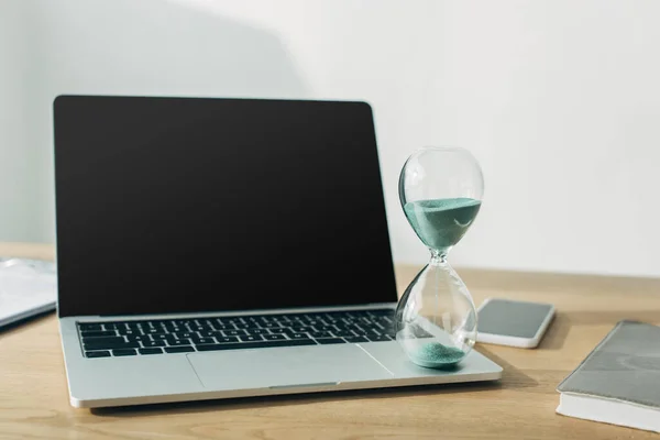 Reloj de arena en el portátil con pantalla en blanco cerca de teléfono inteligente y portátil en la mesa en la oficina - foto de stock