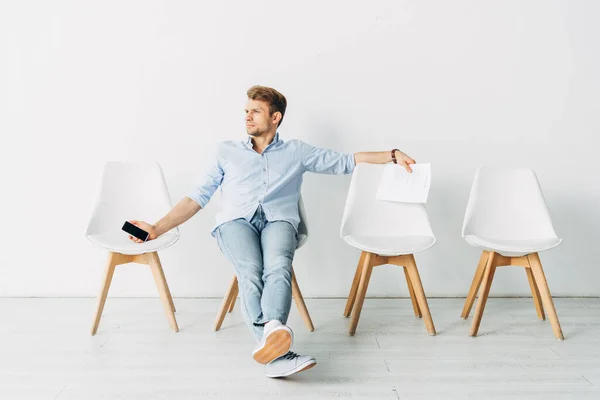 Handsome employee with smartphone and resume sitting on chair in office — Stock Photo