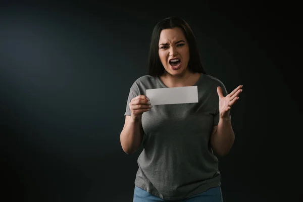 Vista frontal de chica de tamaño grande gritando y sosteniendo la tarjeta vacía sobre fondo negro - foto de stock