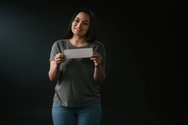 Vista frontal da menina positiva plus size segurando cartão vazio no fundo preto — Fotografia de Stock