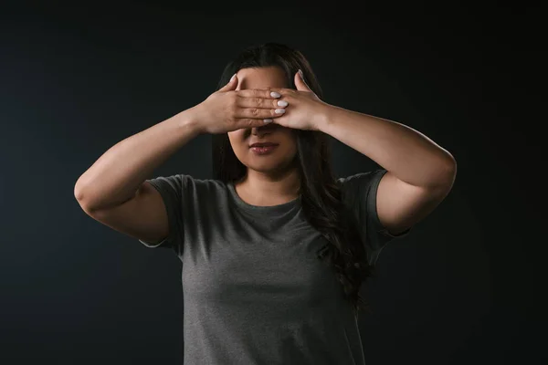 Vista frontal de chica de tamaño grande cubriendo los ojos con las manos sobre fondo negro - foto de stock