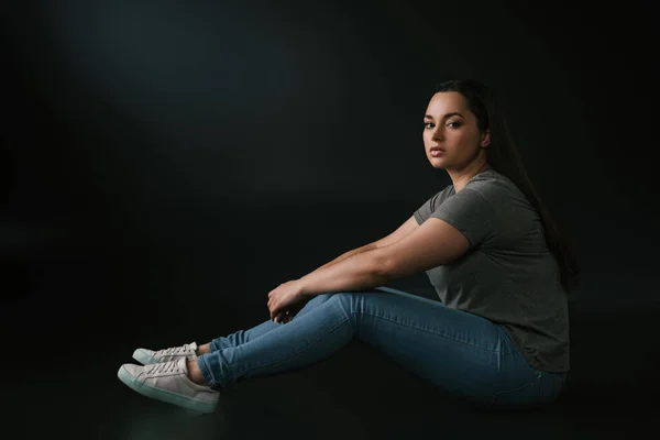 Full length view of beautiful brunette plus size girl sitting on black background — Stock Photo