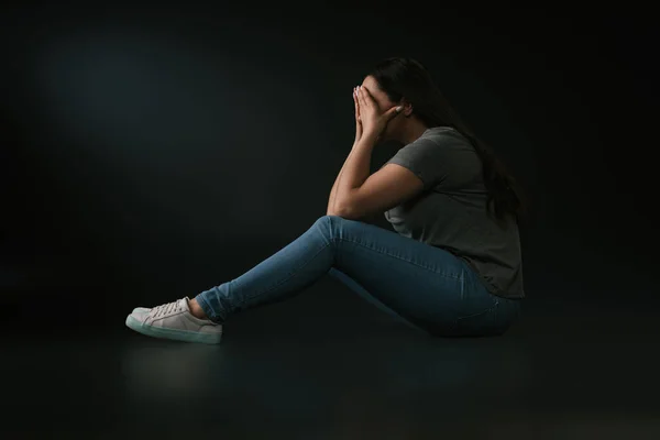 Seitenansicht des weinenden Plus-Size-Mädchens, das Gesicht mit Händen bedeckt und auf schwarzem Hintergrund sitzt — Stockfoto