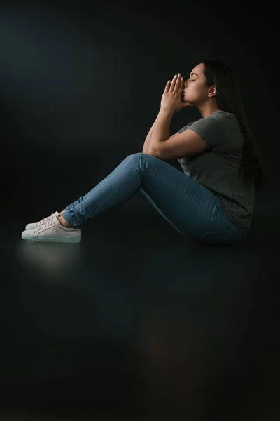 Side view of upset plus size girl covering face with hands and sitting on black background — Stock Photo