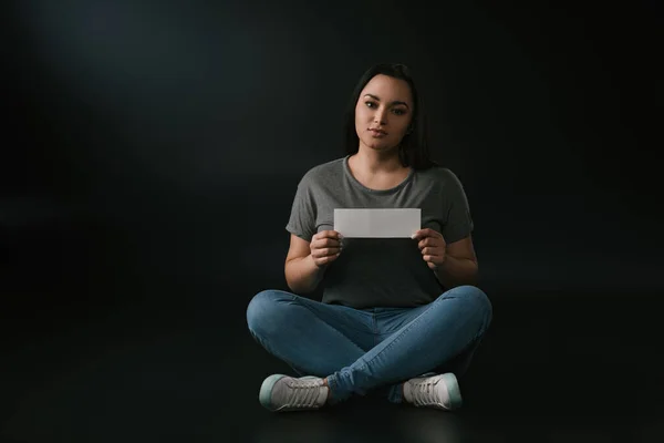 Front view of peaceful plus size girl holding empty card and sitting on black background — Stock Photo