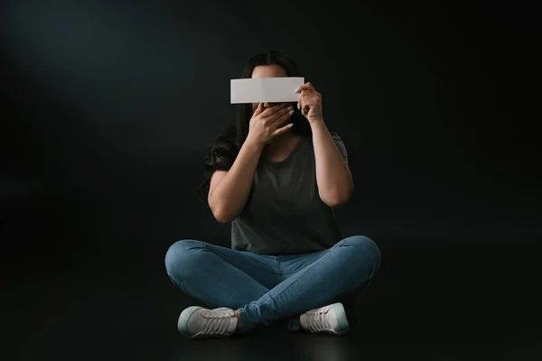 Vista frontal de la chica de tamaño grande que cubre la cara con la mano y la tarjeta vacía y sentado en el fondo negro — Stock Photo