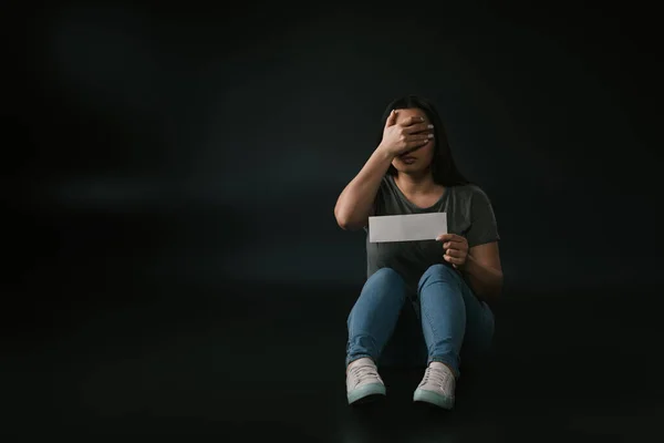 Vista frontal de chica de talla grande con gesto de facepalm y tarjeta vacía en la mano sobre fondo negro - foto de stock